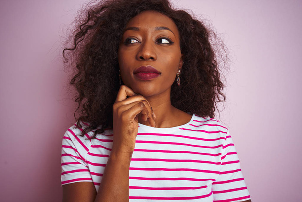 Mladá Afroameričanka v pruhovaném tričku, stojící nad izolovaným růžovým pozadím s rukou na bradě, přemýšlející o otázce a zamyšleném výrazu. Usmíval se zamyšlenou tváří. Pochybný koncept. - Fotografie, Obrázek