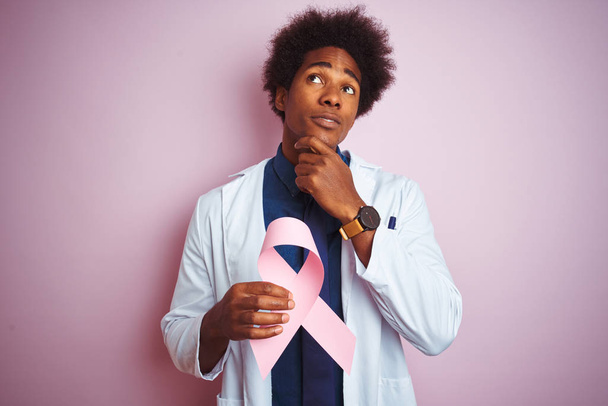 Młody afro amerykański lekarz trzymający wstążkę raka stojącą nad odizolowanym różowym tłem poważna twarz myśląca o pytaniu, bardzo zdezorientowany pomysł - Zdjęcie, obraz