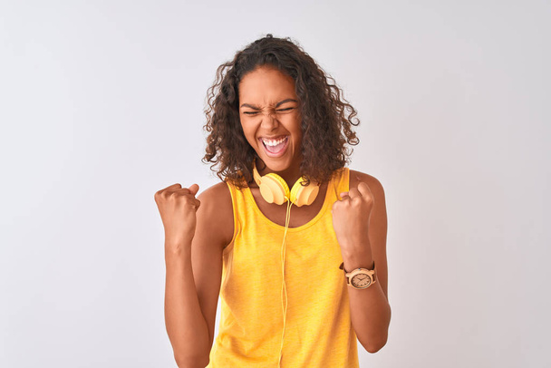 Молодая бразильская женщина в жёлтых наушниках на изолированном белом фоне очень счастлива и взволнована, делая жест победителя с поднятыми руками, улыбаясь и крича об успехе. Концепция празднования
. - Фото, изображение