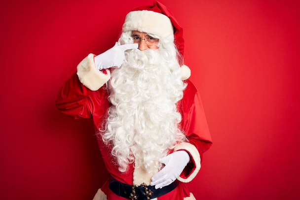 Keski-ikäinen komea mies yllään Santa puku seisoo yli eristetty punainen tausta osoittaa käsin sormella kasvoihin ja nenään, hymyillen iloinen. Kauneus käsite
 - Valokuva, kuva