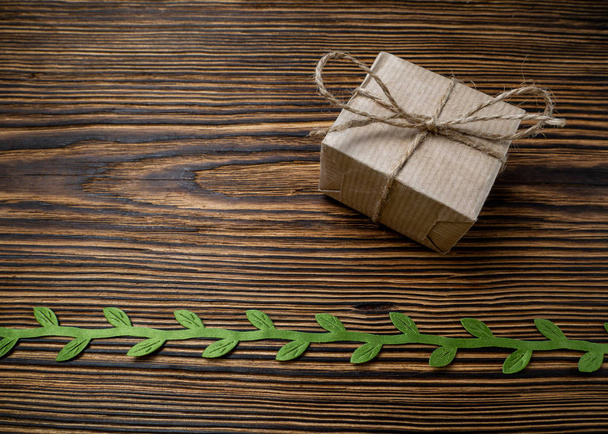 Cadeau. Een doos kraftpapier gebonden met jute touw op een oude, houten achtergrond versierd met groen lint met bladeren - Foto, afbeelding
