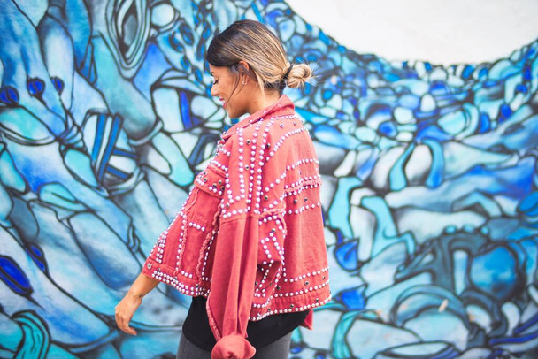 Young beautiful girl wearing red jacket standing backwards over colorful wall  at the town street - Photo, Image