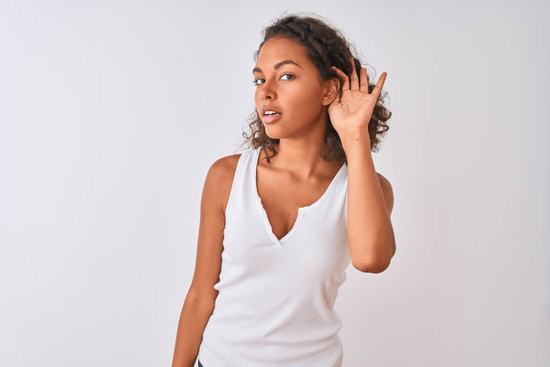 Young brazilian woman wearing casual t-shirt standing over isolated white background smiling with hand over ear listening an hearing to rumor or gossip. Deafness concept. - 写真・画像
