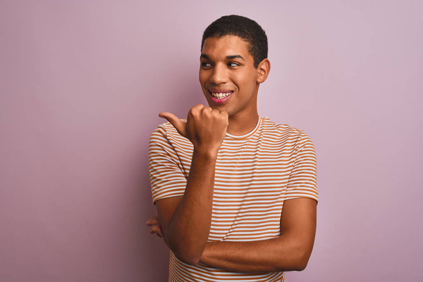Young handsome arab man wearing striped t-shirt standing over isolated pink background smiling with happy face looking and pointing to the side with thumb up. - Photo, image