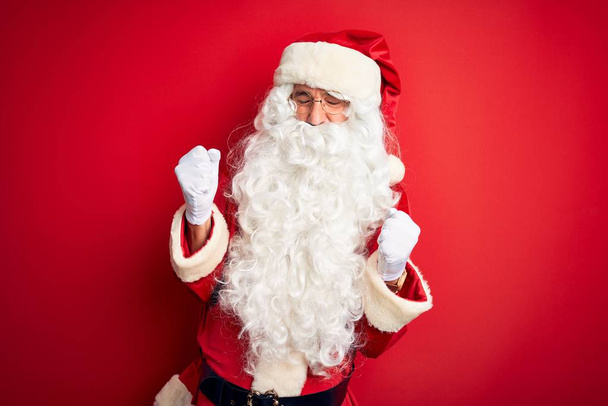 Keski-ikä komea mies yllään Santa puku seisoo yli eristetty punainen tausta erittäin onnellinen ja innoissaan tekee voittaja ele kädet ylhäällä, hymyillen ja huutaen menestystä. Juhlan käsite
. - Valokuva, kuva