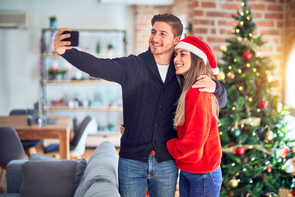 Молодая красивая пара улыбается счастливой и уверенной. Стоя и обнимаясь, сделай селфи на камеру вокруг рождественской елки дома
 - Фото, изображение