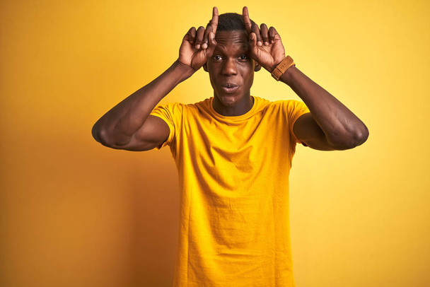 Mladý africký Američan muž na sobě ležérní tričko stojící nad izolovaným žlutým pozadím dělá legrační gesto s prstem přes hlavu jako býčí rohy - Fotografie, Obrázek