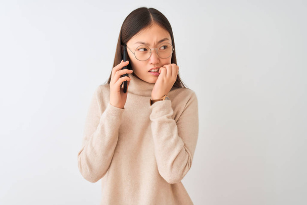 Молода жінка, яка розмовляє на смартфоні через ізольований білий фон, виглядає напруженою і нервовою, з руками кусаючими нігтями. Проблема тривоги. - Фото, зображення