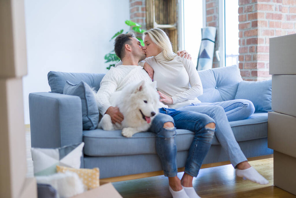 Young beautiful couple with dog kissing sitting on the sofa at new home around cardboard boxes - Fotó, kép