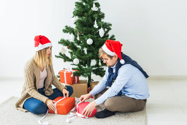 Концепция Рождества и праздников - Молодая счастливая пара в шапках Санты, открывающая подарки дома
. - Фото, изображение
