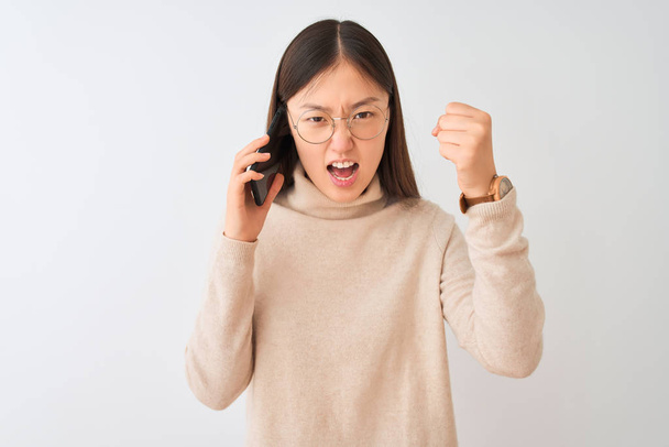Young chinese woman talking on the smartphone over isolated white background angry and mad raising fist frustrated and furious while shouting with anger. Rage and aggressive concept. - Fotografie, Obrázek