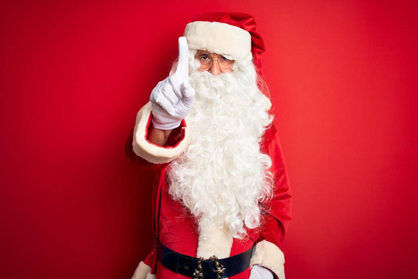 gutaussehender Mann mittleren Alters im Weihnachtsmannkostüm, der mit erhobenem Zeigefinger und wütendem Gesichtsausdruck vor isoliertem rotem Hintergrund steht und keine Geste zeigt - Foto, Bild
