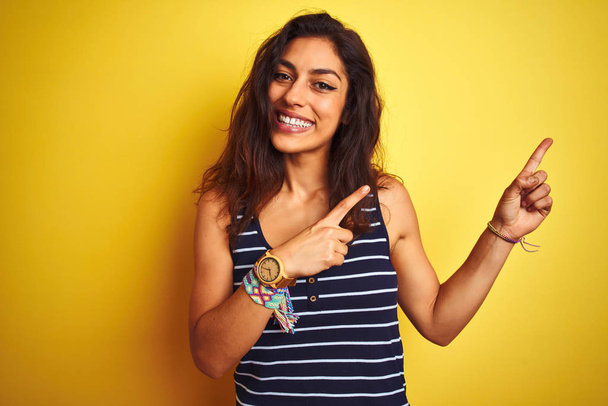 Young beautiful woman wearing striped t-shirt standing over isolated yellow background smiling and looking at the camera pointing with two hands and fingers to the side. - Photo, Image