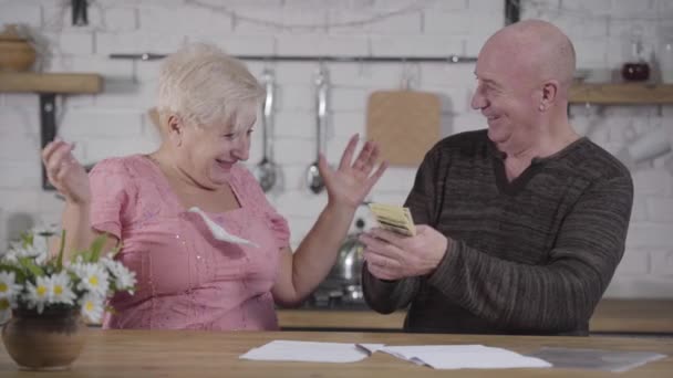 Smiling senior Caucasian man showing pack of banknotes to his shocked wife. Mature woman throwing out bills, taking money and hugging her husband. Joyful elderly couple settling their expenses. - Séquence, vidéo