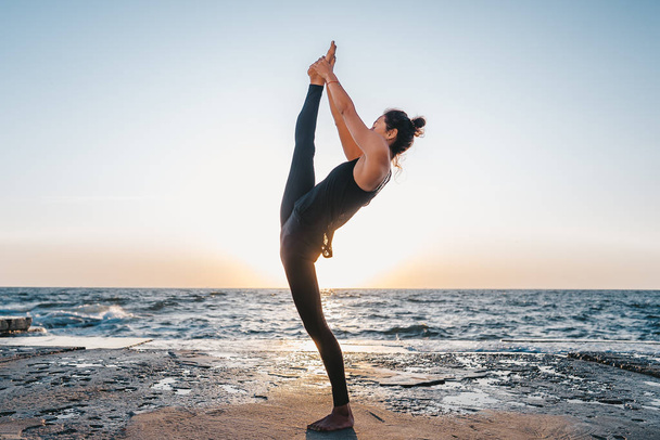 Young woman in black costume practicing yoga at sunrise light, minimalist scene - Foto, immagini