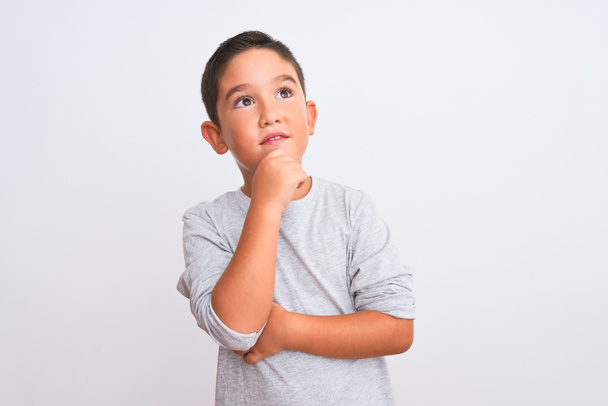 Beautiful kid boy wearing grey casual t-shirt standing over isolated white background with hand on chin thinking about question, pensive expression. Smiling with thoughtful face. Doubt concept. - Photo, Image
