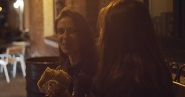 Šťastné ženy přátelé tráví dobrý čas spolu jíst pouliční jídlo venku zpomalený film. Dvě mladé dívky se smějí v městské kavárně a drží cheeseburger s kávou na žluté světlo. Nezdravé dietní jídlo - Záběry, video