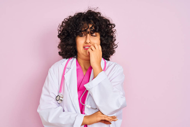 Młoda arabska lekarka z kręconymi włosami, nosząca stetoskop na odizolowanym różowym tle, wyglądająca na zestresowaną i zdenerwowaną dłońmi obgryzającymi paznokcie. Problem lękowy. - Zdjęcie, obraz