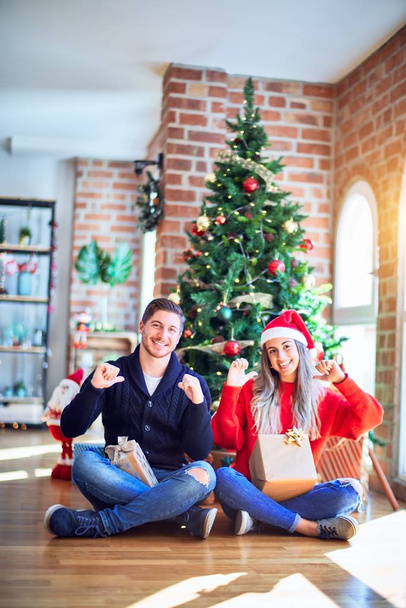 Молодая пара в шляпе Санта-Клауса сидит на полу вокруг рождественской елки дома и смотрит уверенно с улыбкой на лице, указывая на себя пальцами гордым и счастливым
. - Фото, изображение