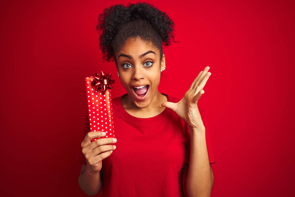 Joven mujer afroamericana sosteniendo regalo de San Valentín sobre fondo rojo aislado muy feliz y emocionado, expresión ganadora celebrando la victoria gritando con una gran sonrisa y las manos levantadas
 - Foto, imagen