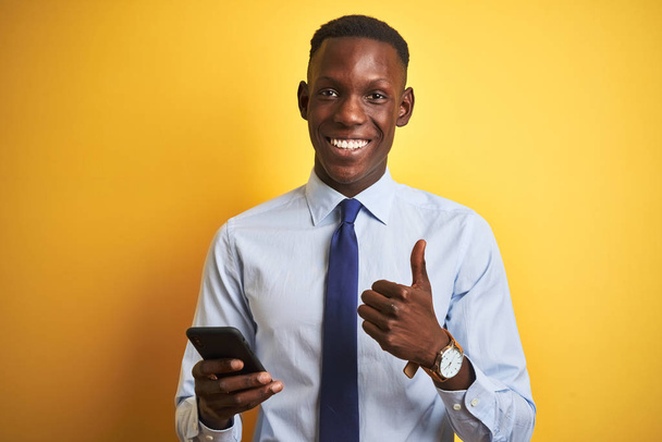 Empresário afro-americano usando smartphone em pé sobre fundo amarelo isolado feliz com grande sorriso fazendo sinal ok, polegar com os dedos, excelente sinal
 - Foto, Imagem