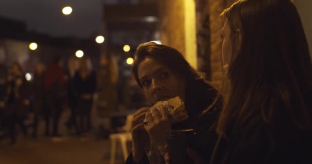 Dvě hladové dívky jedí fast food svačinu sedí na ulici města ve večerním zpomaleném filmu. Šťastné přítelkyně tráví volný čas spolu venku na městském nočním pozadí. Mládež bezstarostný životní styl - Záběry, video