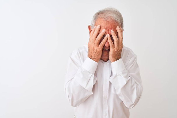 Starszy siwowłosy mężczyzna ubrany w elegancką koszulę stojący nad odizolowanym białym tłem ze smutnym wyrazem twarzy pokrywającym dłonie podczas płaczu. Koncepcja depresji. - Zdjęcie, obraz