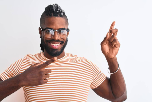 Afro-Amerikaanse man draagt gestreept t-shirt en bril over geïsoleerde witte achtergrond glimlachend en kijkend naar de camera wijzend met twee handen en vingers naar de zijkant. - Foto, afbeelding