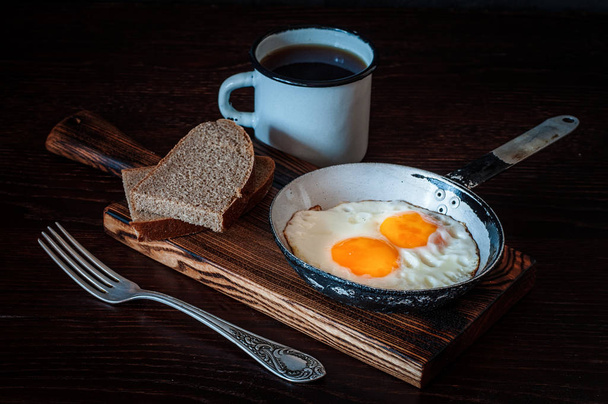 Fried scrambled eggs in vintage pan, with bread, old fork, metal mug with tea, top view - Photo, Image
