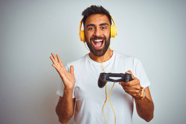 Arab indian gamer man playing video game using headphones over isolated white background very happy and excited, winner expression celebrating victory screaming with big smile and raised hands - Photo, image