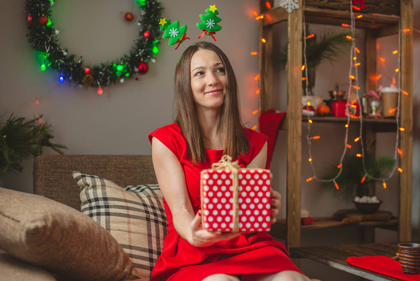 Frau in rotem Kleid sitzt auf einem Sofa mit einer Geschenkschachtel in der Hand vor dem Hintergrund von Lichterketten. Gemütliche Weihnachtsstimmung - Foto, Bild