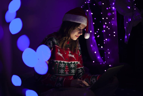 young girl with a laptop in the New Year sits at home in Christmas clothes, a woman online for Christmas - Foto, Imagen