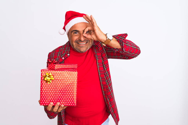 Middle age man wearing Christmas Santa hat holding gift over isolated white background with happy face smiling doing ok sign with hand on eye looking through fingers - Фото, зображення