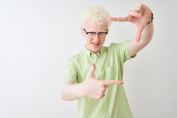 Young albino blond man wearing green shirt and glasses over isolated white background smiling making frame with hands and fingers with happy face. Creativity and photography concept. - Photo, Image