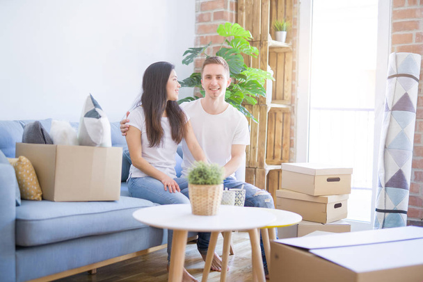 Young beautiful couple sitting on the sofa drinking coffee at new home around cardboard boxes - Photo, Image