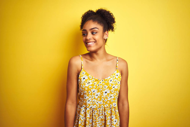 African american woman wearing casual floral dress standing over isolated yellow background looking away to side with smile on face, natural expression. Laughing confident. - Valokuva, kuva