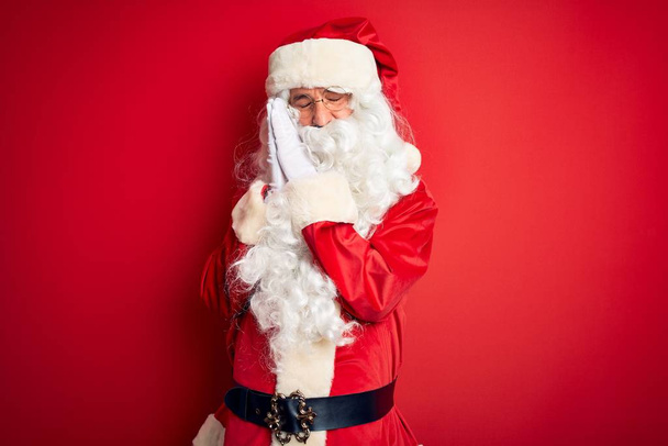 gutaussehender Mann mittleren Alters im Weihnachtsmannkostüm, der vor isoliertem roten Hintergrund steht und müde träumt und mit geschlossenen Augen lächelnd mit den Händen posiert. - Foto, Bild