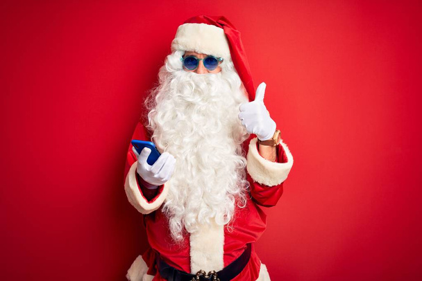 Hombre mayor con disfraz de Papá Noel usando un teléfono inteligente sobre un fondo rojo aislado feliz con una gran sonrisa haciendo buen signo, pulgar hacia arriba con los dedos, excelente signo
 - Foto, imagen