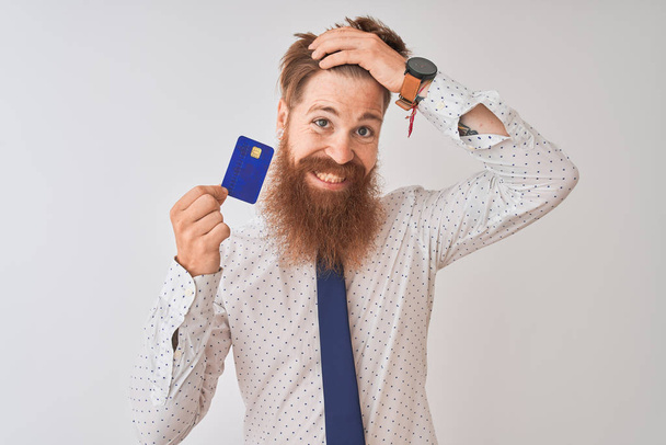 Jeune homme d'affaires irlandais roux tenant la carte de crédit debout sur fond blanc isolé stressé avec la main sur la tête, choqué par la honte et le visage surprise, en colère et frustré. Peur et contrariété par erreur
. - Photo, image