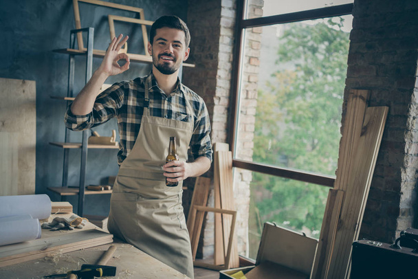 Photo of cheerful bearded positive man showing ok sign in apron checkered shirt holding bottle of beer having finished woodwork - Fotografie, Obrázek