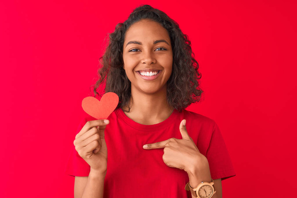 Giovane donna brasiliana che tiene il cuore di carta in piedi sopra isolato sfondo rosso con faccia a sorpresa puntando il dito verso se stesso
 - Foto, immagini