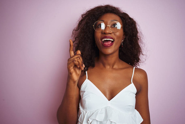 Mujer afroamericana joven con gafas de pie sobre un fondo rosa aislado apuntando con el dedo hacia arriba con una idea exitosa. Salido y feliz. Número uno.
. - Foto, imagen