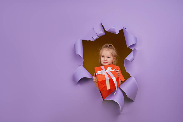 Schattig klein meisje breekt door een gekleurde paarse papieren muur met geschenkdoos in handen. Peuter grappige emoties gezicht. Kopieer ruimte voor tekst - Foto, afbeelding