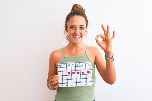 Jeune rousse femme tenant calendrier menstruation sur fond isolé faisant signe ok avec les doigts, excellent symbole
 - Photo, image