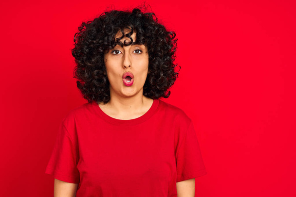 Young arab woman with curly hair wearing casual t-shirt over isolated red background afraid and shocked with surprise expression, fear and excited face. - Φωτογραφία, εικόνα