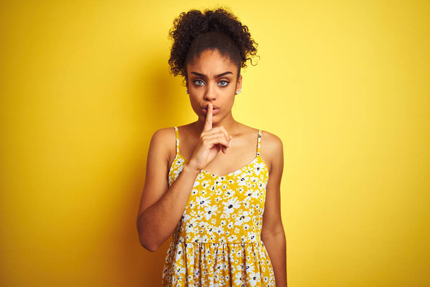 Femme afro-américaine portant une robe florale décontractée debout sur un fond jaune isolé demandant à être tranquille avec le doigt sur les lèvres. Silence et concept secret
. - Photo, image