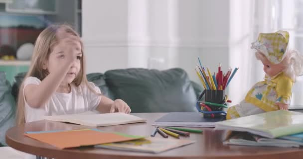 Concentrated Caucasian schoolgirl explaining math to the doll placed on the table. Clever pretty girl doing homework at home. Fun in education, intelligence, studying concept. Cinema 4k ProRes HQ. - Кадри, відео