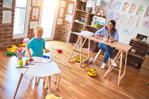 Young caucasian child playing at playschool with teacher. Young woman sitting on the desk of the classroom - Photo, Image