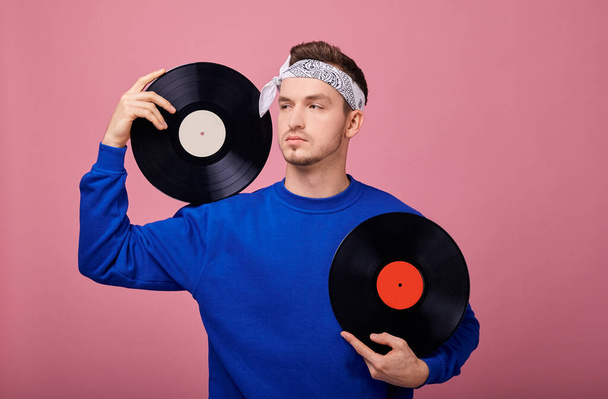 Close-up. A cool stylish guy in a bandana in a dark blue jacket stands on a pink background with a black vinyl record in his hand. Youth, style, joy, playful mood. Retro style. - Foto, imagen