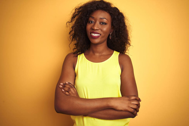 Mladá Afroameričanka v tričku, stojící nad osamělým žlutým pozadím, veselá tvář se zkříženýma rukama, dívající se do kamery. Pozitivní osoba. - Fotografie, Obrázek